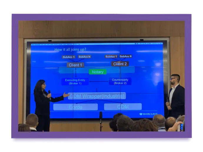 Two finance professionals presenting a flow chart on a large screen titled "How it all joins up?", illustrating the integration of CDM, Corda, Clients, and a Notary in a financial technology context, likely related to distributed ledger or blockchain technology at a Barclays event.